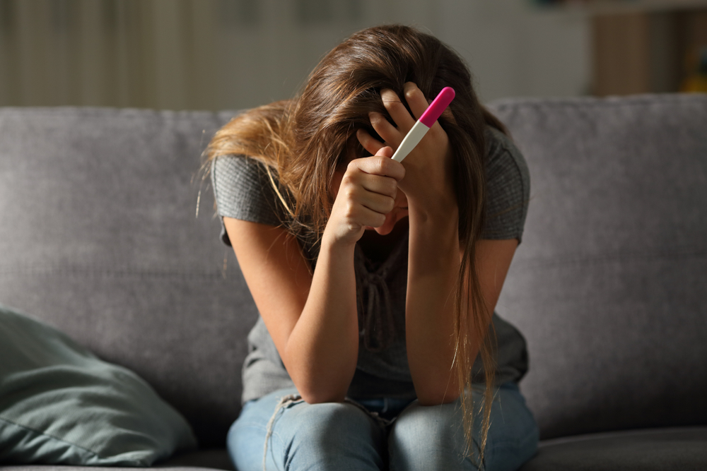 sad woman sitting alone holding a pregnancy test sitting on a couch in the living room at home