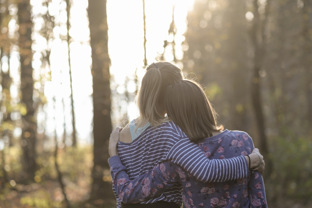 View from behind of two friends standing in autumn woods leaning on each other with their arms around one another.